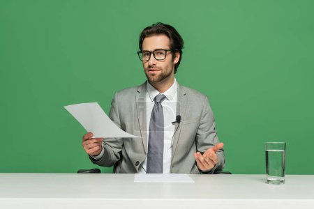 Photo for News broadcaster in eyeglasses and suit sitting at desk and holding paper isolated on green - Royalty Free Image