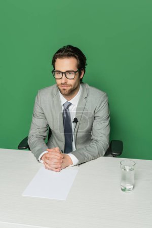 overhead view of news broadcaster in eyeglasses and suit sitting with clenched hands at desk isolated on green