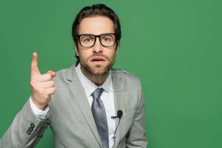 emotional newscaster in eyeglasses and suit pointing with finger isolated on green 