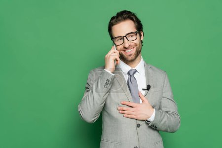fröhlicher Reporter in Brille und Anzug, der auf grünem Hintergrund mit dem Smartphone spricht 