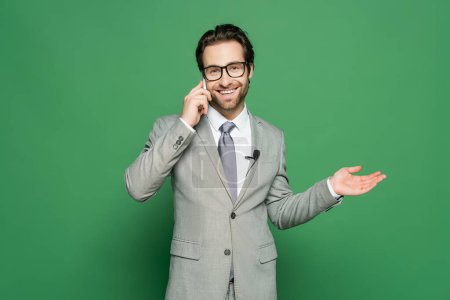 Lächelnder Reporter in Brille und Anzug spricht auf Smartphone auf grünem Hintergrund 