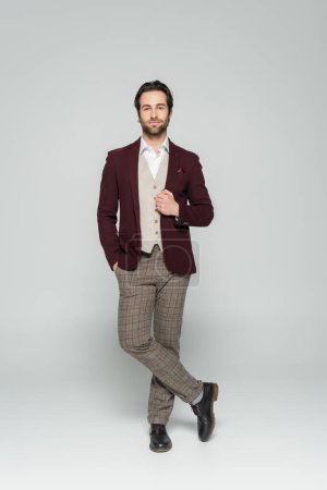 longitud completa de showman barbudo en chaleco y chaqueta roja de pie con la mano en el bolsillo en gris  