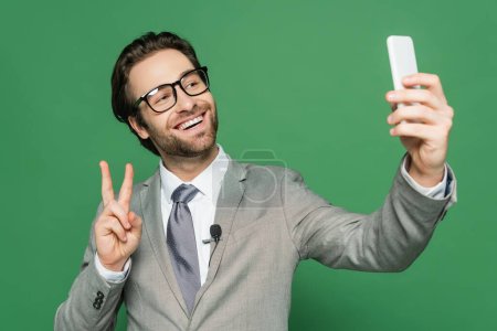 Lächelnder Reporter in Brille und Anzug, der ein Selfie auf dem Smartphone macht 