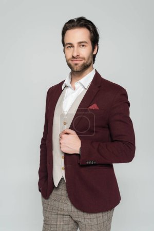 bearded showman in vest and red blazer standing with hand in pocket isolated on grey 