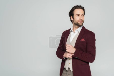 Foto de Showman barbudo en chaleco y chaqueta roja de pie y mirando hacia otro lado aislado en gris - Imagen libre de derechos
