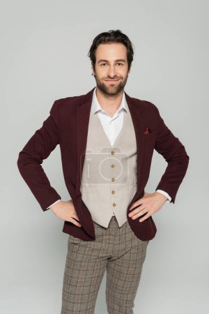 cheerful showman in vest and red blazer standing with hands on hips isolated on grey 