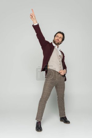 Foto de Longitud completa de showman barbudo en chaleco y chaqueta roja de pie con la mano levantada en gris - Imagen libre de derechos