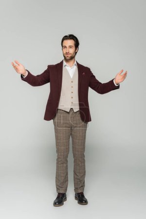 Photo for Full length of bearded host of event in vest and red blazer standing with outstretched hands on grey - Royalty Free Image