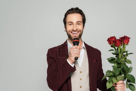 cheerful host of event in blazer holding red roses and talking in microphone isolated on grey 