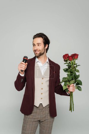 Photo for Bearded host of event in blazer holding red roses and talking in microphone isolated on grey - Royalty Free Image