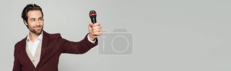 Photo for Cheerful host of event in formal wear looking at microphone isolated on grey, banner - Royalty Free Image