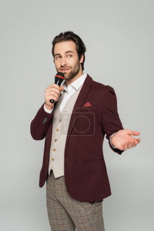 bearded host of event in formal wear making announcement in microphone and gesturing isolated on grey 