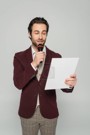 bärtige Moderatorin in formeller Kleidung, die Ansage in Mikrofon macht, während sie aus Papier isoliert auf grau liest 