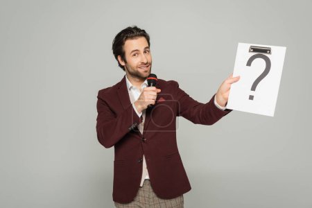 bearded host of event in formal wear talking in microphone while holding clipboard with question mark isolated on grey 