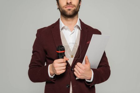 cropped view of bearded host of event in formal wear holding microphone and clipboard isolated on grey 