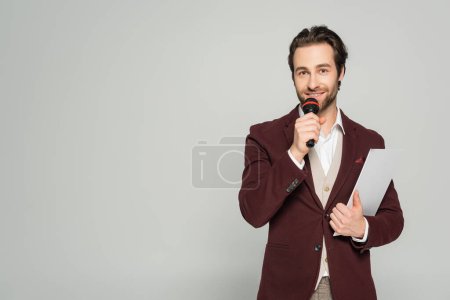Foto de Alegre anfitrión de evento en ropa formal hablando en micrófono y sujetando portapapeles aislado en gris - Imagen libre de derechos