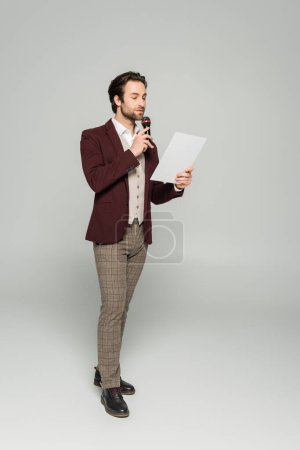 Foto de Longitud completa de showman barbudo en ropa formal hablando en micrófono y mirando el papel en gris - Imagen libre de derechos