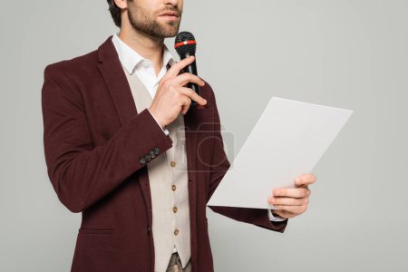cropped view of bearded showman in formal wear talking in microphone and holding paper on grey 