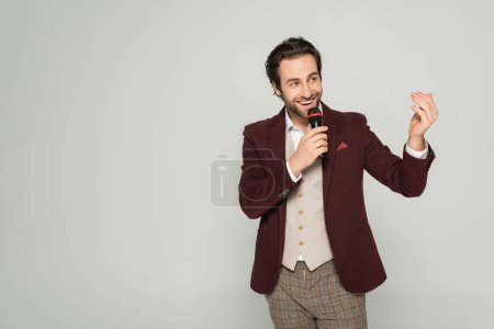 happy entertainer in formal wear talking in microphone and looking away isolated on grey 
