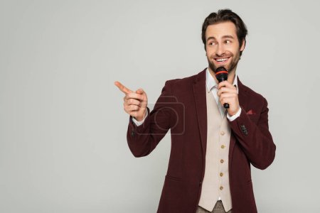 positive showman in formal wear talking in microphone and pointing with finger isolated on grey 