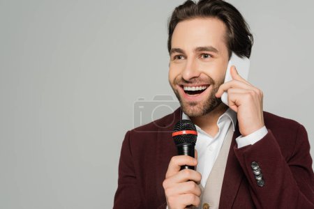 fröhlicher Showman, der auf dem Smartphone spricht, während er das Mikrofon isoliert auf grau hält 