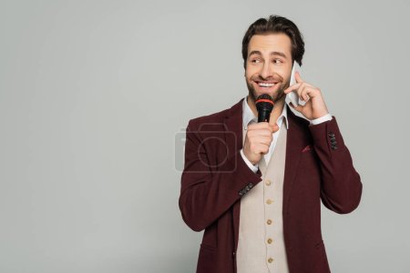 happy showman talking on smartphone while holding microphone isolated on grey 