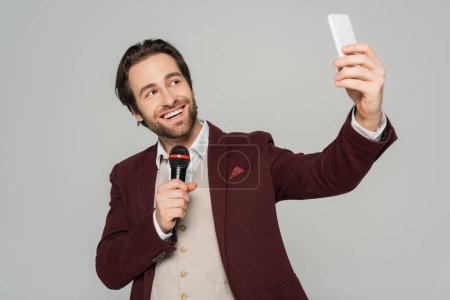 fröhlicher Showman macht Selfie mit Smartphone und hält Mikrofon isoliert auf grau 