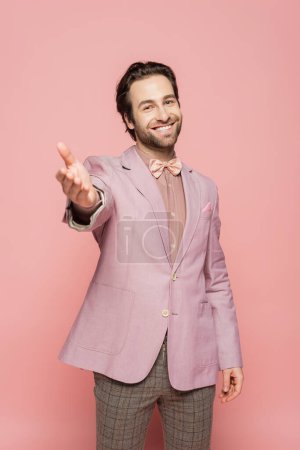 cheerful and young showman in suit with bow tie standing with outstretched hand isolated on pink 