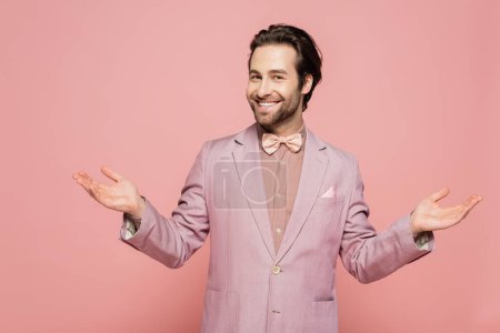 fröhlicher und junger Showman im Anzug mit Fliege gestikuliert vereinzelt auf rosa 