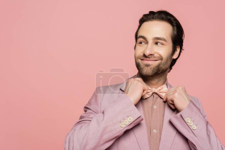positive and young showman in suit adjusting bow tie isolated on pink 