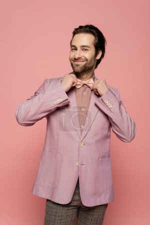 cheerful and young showman in suit adjusting bow tie isolated on pink 