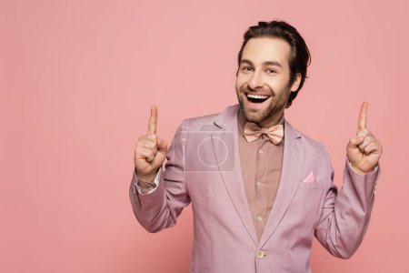 fröhlicher und junger Showman im Anzug mit Fliege, die nach oben zeigt, mit Fingern vereinzelt auf rosa 