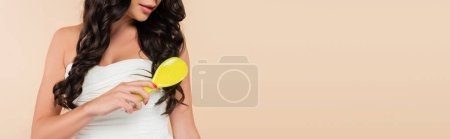 Cropped view of brunette woman combing wavy hair isolated on beige, banner 