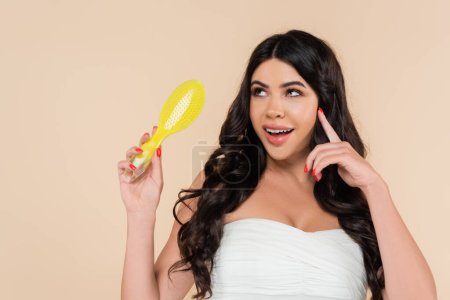 Dreamy woman in top holding hairbrush and looking away isolated on beige 