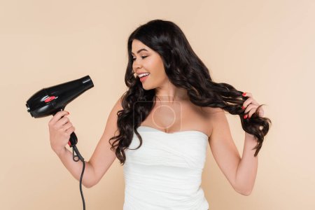 Cheerful brunette woman holding hairdryer and touching hair isolated on beige 
