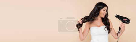 Positive brunette woman drying hair isolated on beige with copy space, banner 