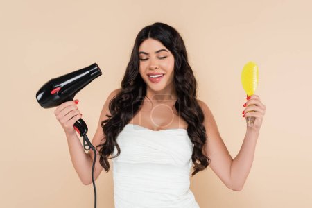 Smiling brunette woman holding hairbrush and hairdryer isolated on beige 