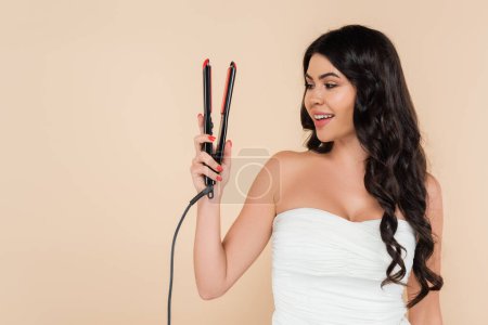 Smiling brunette woman with hairstyle looking at curling iron isolated on beige 