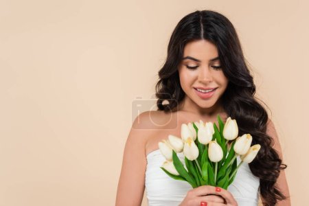 Positive brunette woman looking at white tulips isolated on beige 