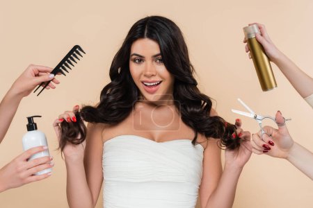 Hands with hairdressing tools near cheerful brunette woman isolated on beige 