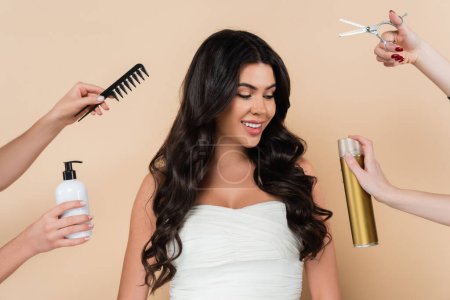 Smiling woman with wavy hairstyle looking at hands holding hairdressing tools isolated on beige 