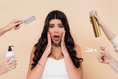Shocked brunette woman looking at camera near hands with hairdressing tools isolated on beige 