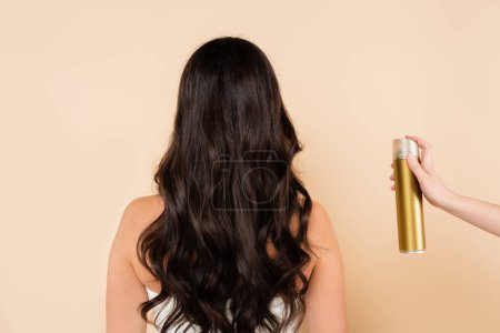 Back view of woman with wavy hair standing near hand with hairspray isolated on beige 