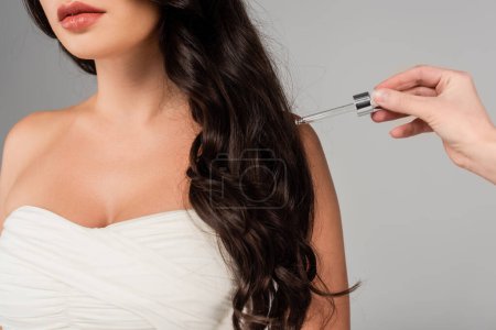 Cropped view of brunette woman standing near hand holding dropper of hair oil isolated on grey 