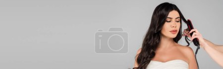 hairdresser styling hair of brunette woman with hair straightener isolated on grey, banner 