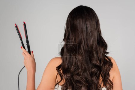 Foto de Back view of curly woman holding hair straightener isolated on grey - Imagen libre de derechos