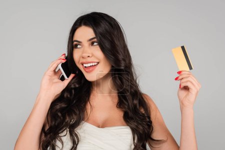 pretty woman with long and shiny hair holding credit card and talking on mobile phone isolated on grey