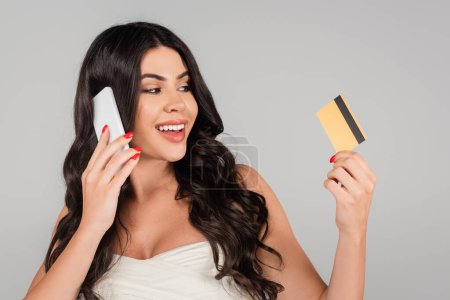 happy brunette woman with long hair holding credit card and talking on smartphone isolated on grey