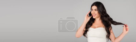 happy brunette woman talking on cellphone while touching healthy hair isolated on grey, banner