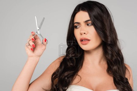 Foto de Worried brunette woman with long and shiny hair holding thinning scissors isolated on grey - Imagen libre de derechos
